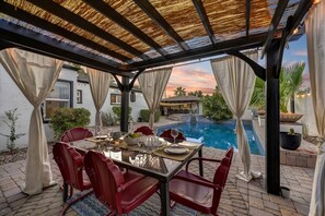 Eine Außenpatio mit Pool, roten Stühlen, einem Essplatz und einer Pergola mit einem Reetdach.
