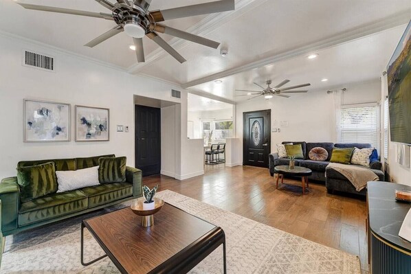 A spacious living room with a green sofa, a wooden coffee table, and a dark armchair. There are ceiling fans and a large window with blinds.
