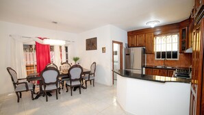 A dining area with a table and chairs, a kitchen with wooden cabinets and a refrigerator, and a window with curtains. - Spacious and secure villa 5 minutes from the beach (Puerto Plata)