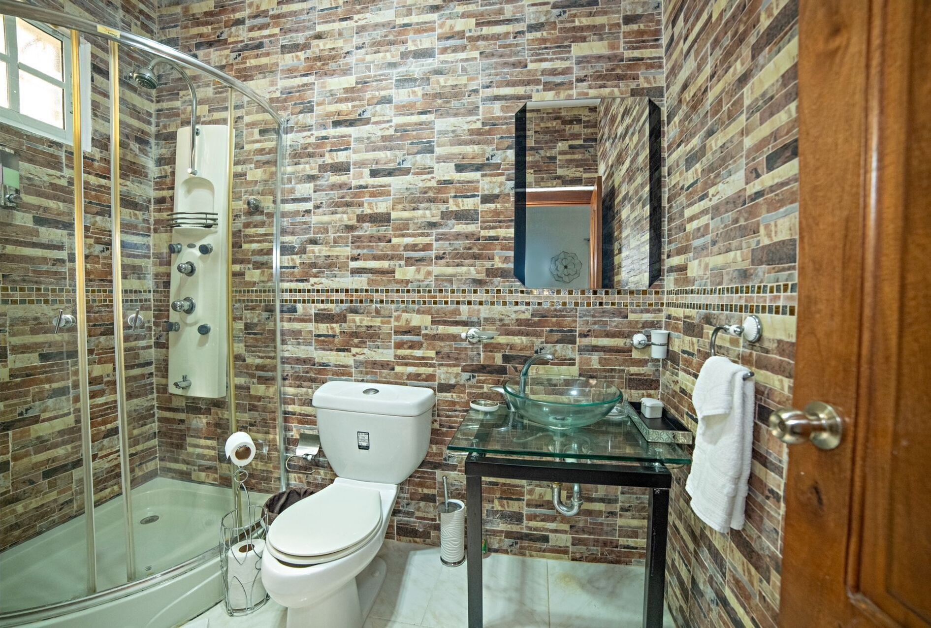 A bathroom with a glass shower enclosure, a toilet, a glass sink, and a mirror on the wall.