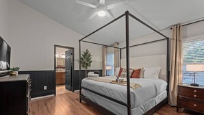 Un lit à baldaquin, une commode en bois, un téléviseur et une plante en pot dans une chambre moderne dotée d’un plancher en bois et d’un ventilateur de plafond.
