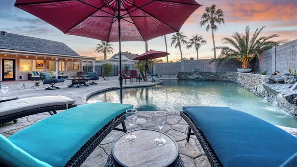 A pool area with lounge chairs, a table with a glass, and umbrellas. - PRIVATE OASIS with the BEST backyard!!! (Phoenix)