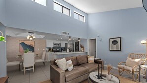 Un salon moderne avec un canapé, une table basse et des fauteuils. Il y a également un coin cuisine avec une table à manger et des chaises. La pièce est dotée d’un haut plafond et de grandes fenêtres.