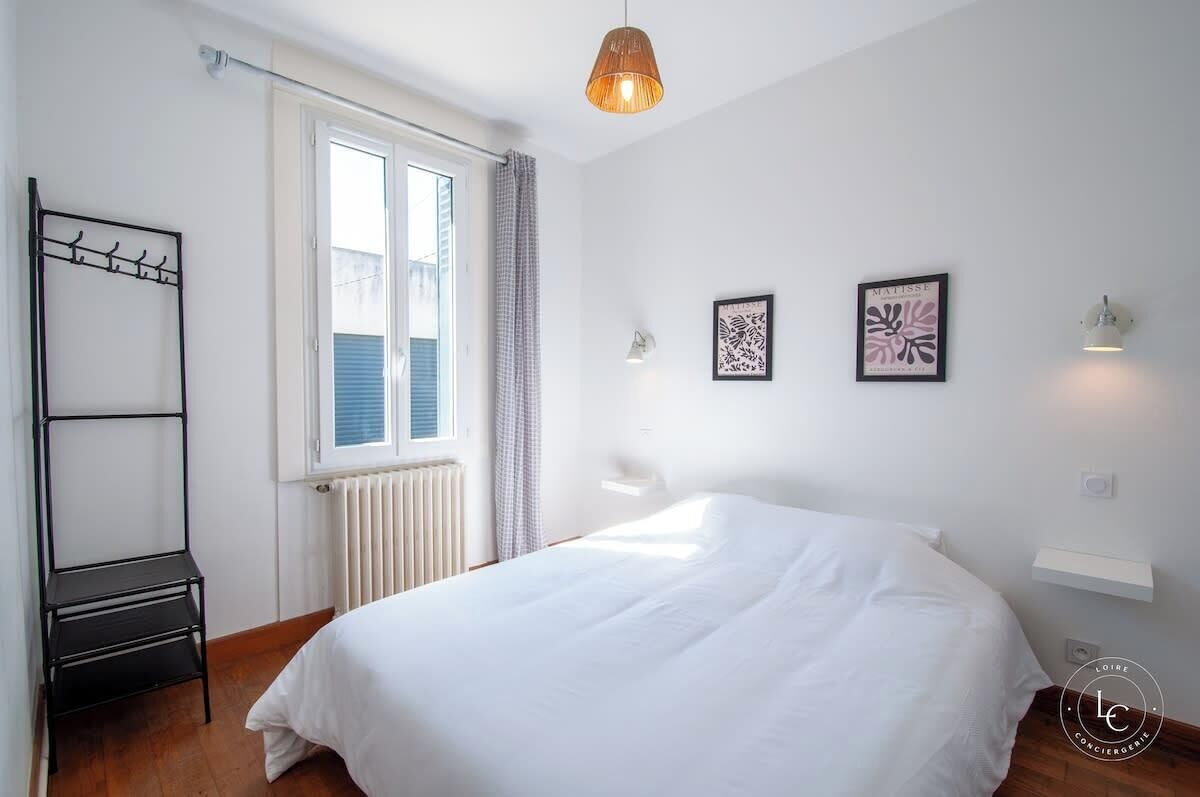 A bedroom with a bed, a radiator, a window with curtains, a wall-mounted shelf, and a hanging light fixture.