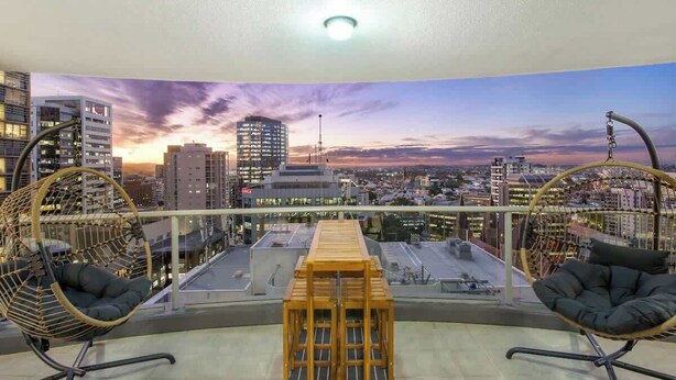 A rooftop terrace with two wicker chairs, a wooden table, and a cityscape view at sunset. - Arcade In The Sky- Views, Games & Vibes (Brisbane City)