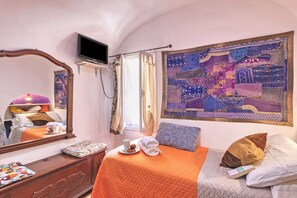 A bedroom with a bed, a television, a window with curtains, a tapestry, and a bedside table with a cup and a book.