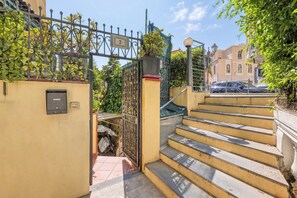 Una entrada con puerta y cerrojo, un letrero con número, una escalera y un edificio al fondo.