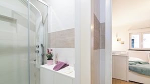 Une salle de bain moderne avec une douche à l’italienne en verre, un meuble-lavabo blanc agrémenté d’une plante rose, et un lit recouvert d’une couverture verte dans la pièce adjacente.