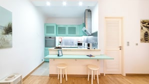 Une cuisine compacte avec des meubles de couleur turquoise, un réfrigérateur en acier inoxydable et un plan de travail blanc. On y trouve deux tabourets blancs et un tableau encadré posé sur le plan de travail.