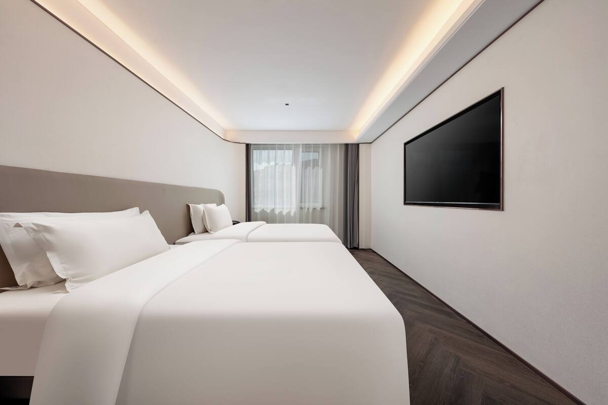 Una habitación de hotel moderna con una cama grande, un televisor de pantalla plana en la pared y un ventanal con cortinas translúcidas.