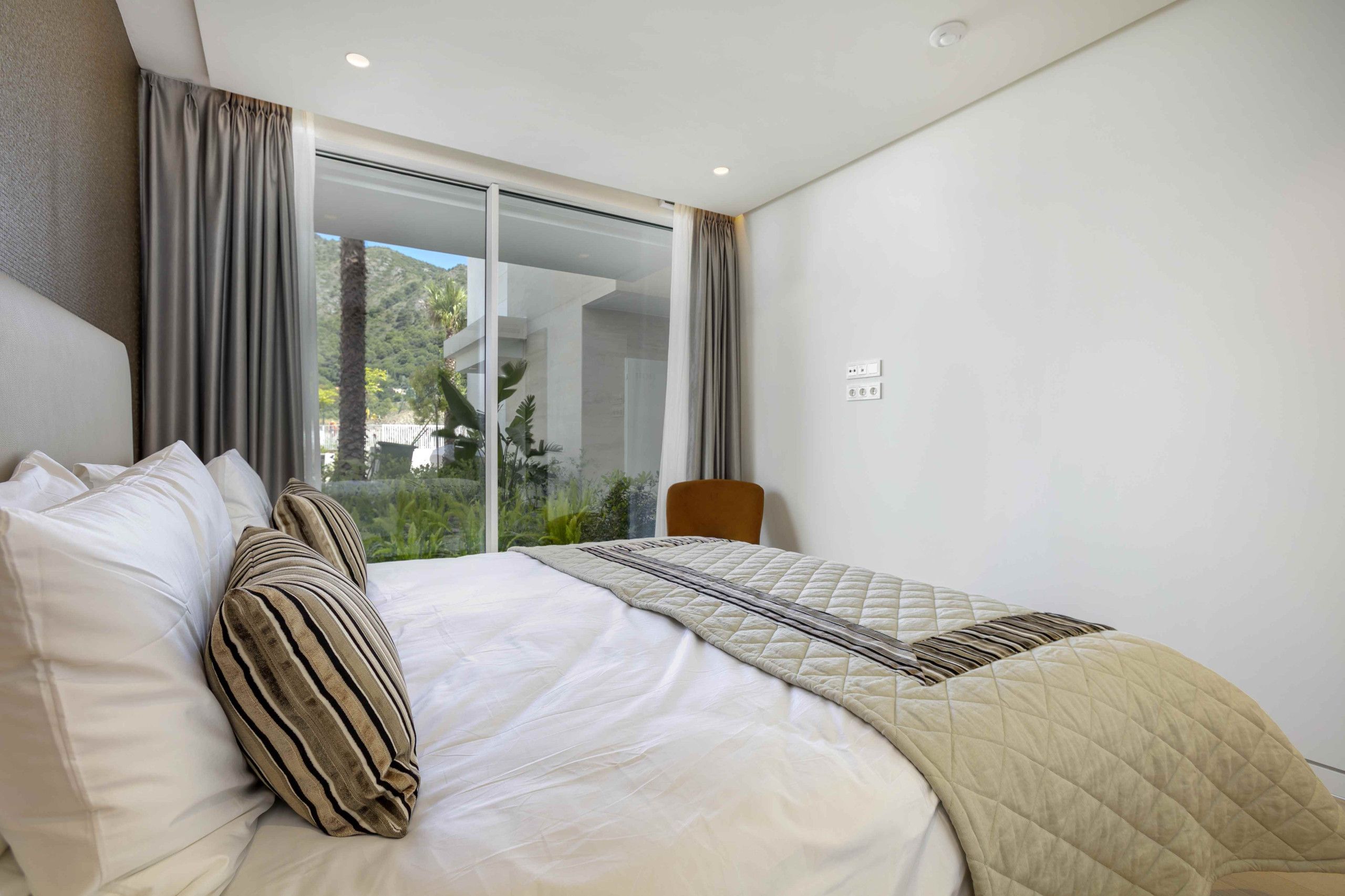 A modern hotel room with a large bed, a view through a sliding glass door, and a chair by the window.