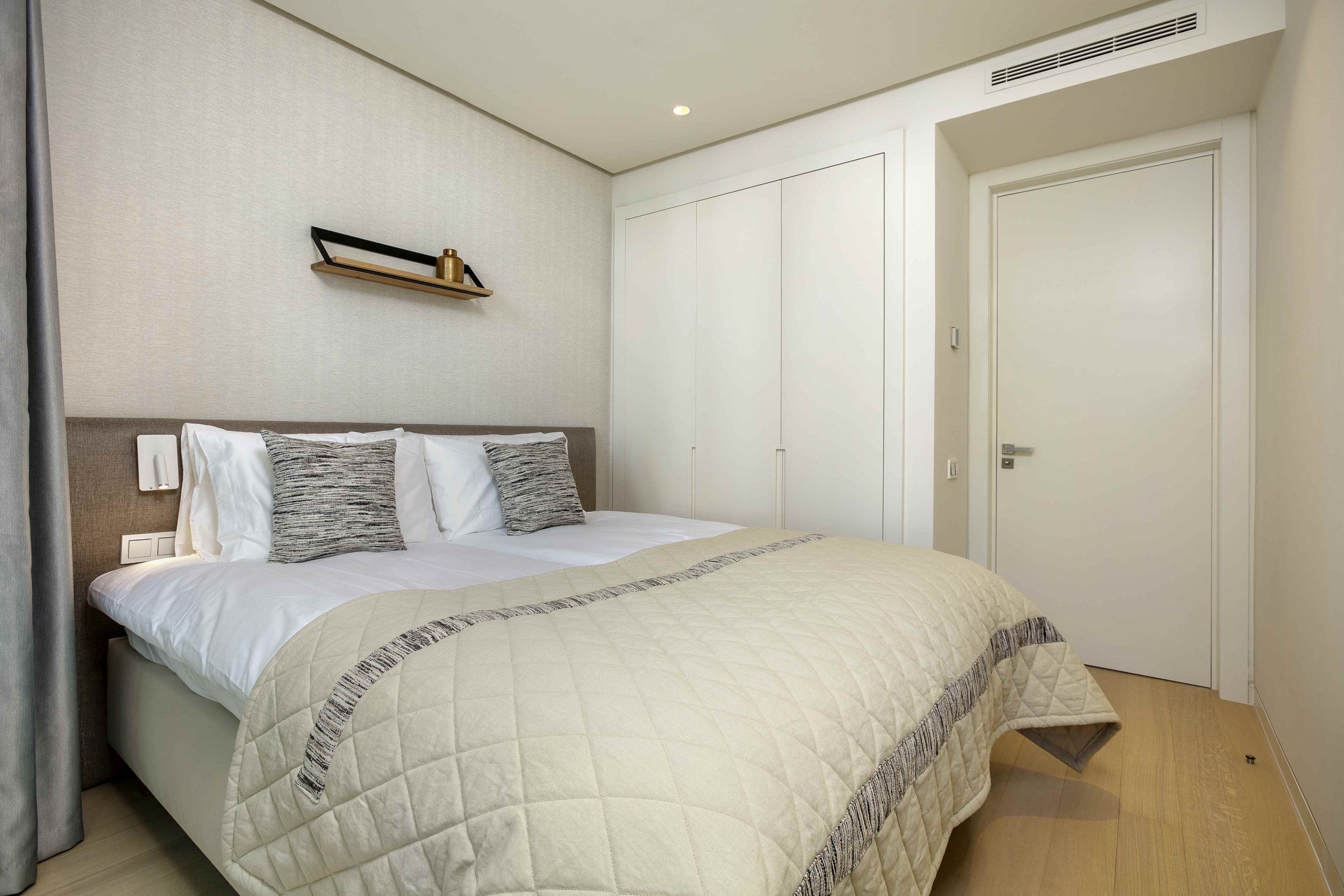 A neatly arranged bedroom with a large bed, a bedside table, a wall-mounted shelf, and a wardrobe.