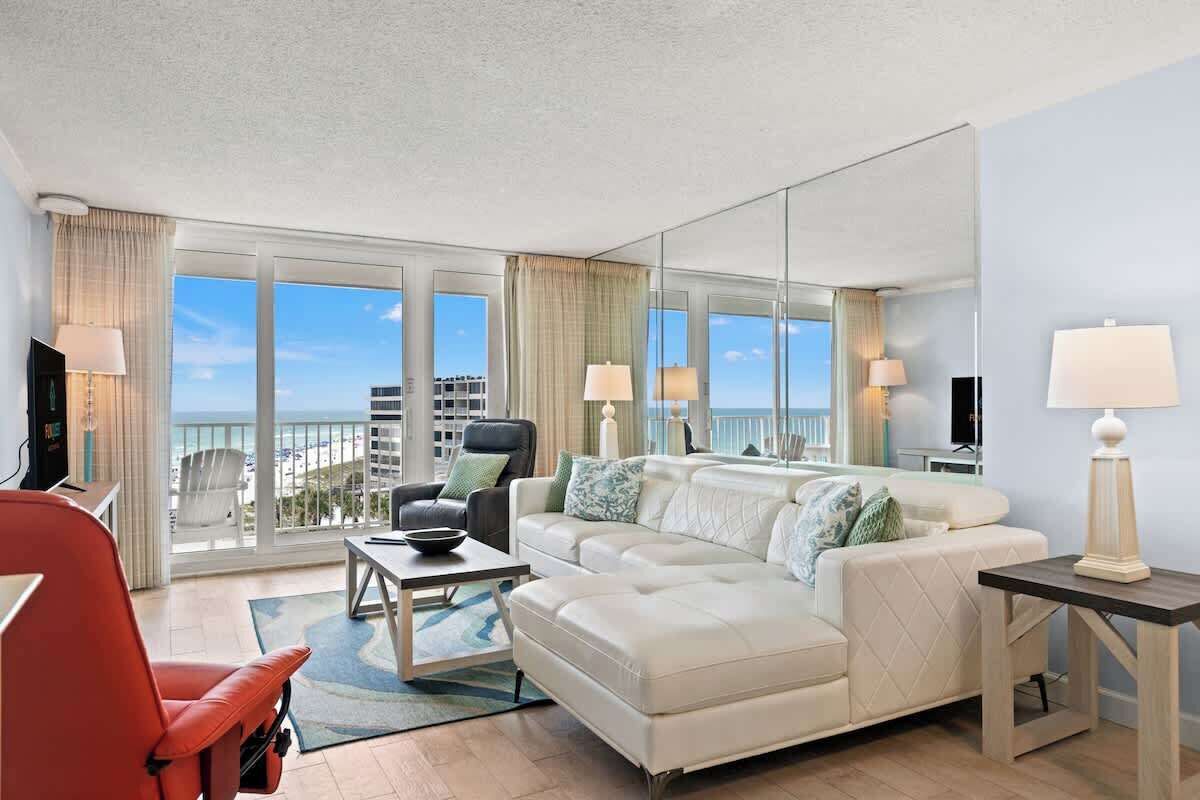 Quarto de hotel moderno com uma janela grande que oferece vista da cidade, um sofá branco, uma cadeira vermelha e uma mesinha com um abajur.