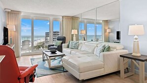 Habitación de hotel moderna con un ventanal amplio que ofrece vistas a la ciudad, un sofá blanco, una silla roja y una mesita con una lámpara.