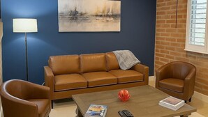 Ein Wohnzimmer mit einem braunen Ledersofa, zwei braunen Ledersesseln, einem Couchtisch mit Zeitschriften und Fernbedienungen, einer Stehlampe und einem Gemälde an der Wand.
