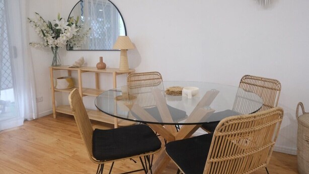 Un comedor con una mesa redonda de vidrio, cuatro sillas de mimbre con cojines negros, un espejo, un florero con flores y una lámpara sobre una estantería de madera.