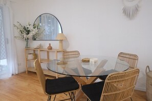 Un coin repas avec une table ronde en verre, quatre chaises en osier avec des coussins noirs, un miroir, un vase avec des fleurs et une lampe posée sur une étagère en bois.