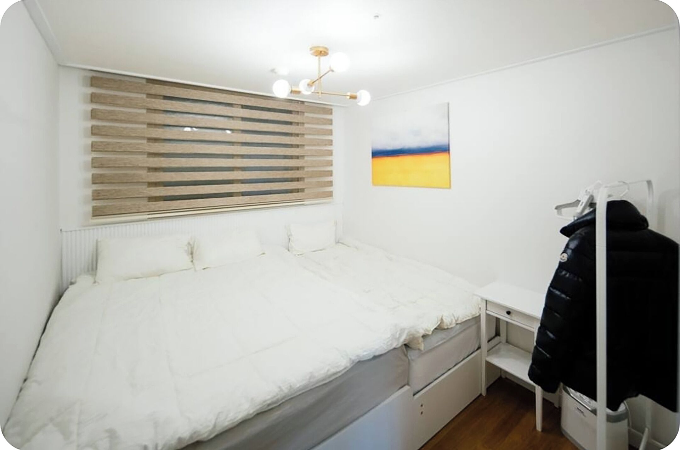 A bedroom with a bed, a desk, a chair, a radiator, a window with blinds, and a painting on the wall.