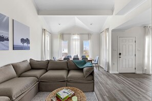 A living room with a sectional sofa, a dining table with chairs, and a door leading to another room.