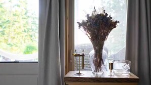 A window with a vase of dried flowers on a wooden table, a candle holder, and a clear glass candle.