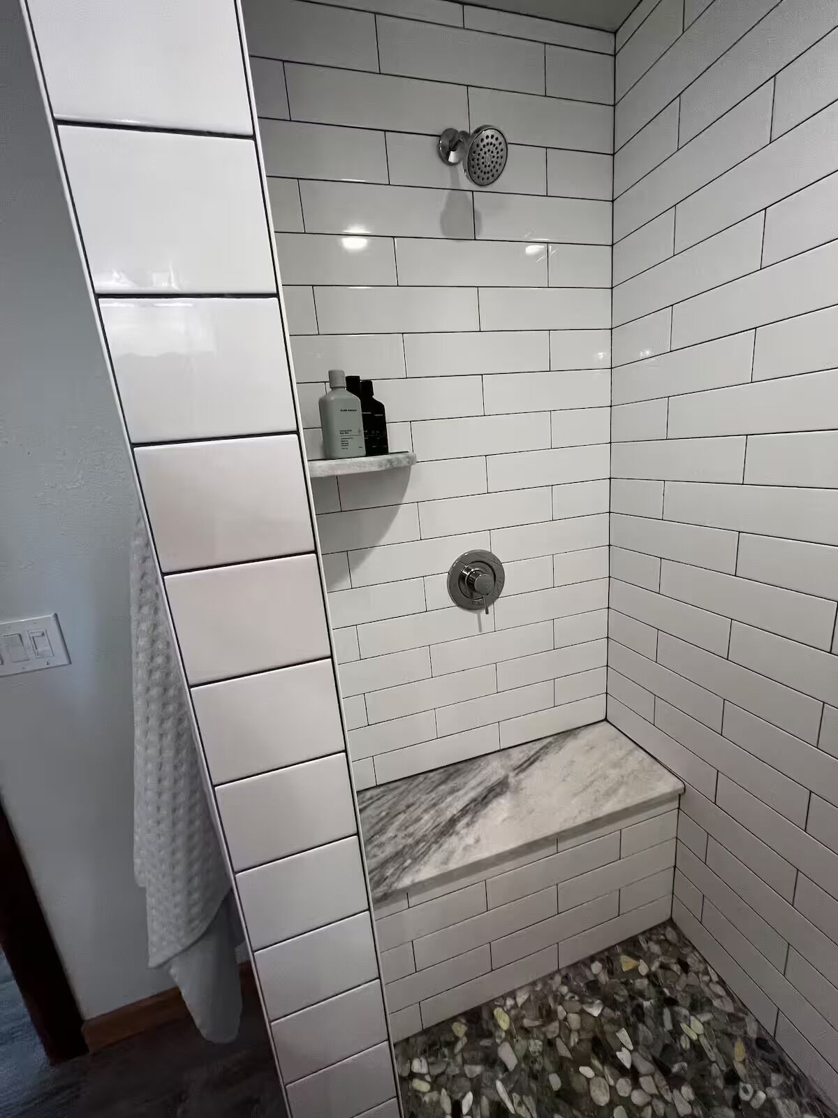 En dusch med vita kakelplattor, en bänk i marmor och en hylla med toalettartiklar.