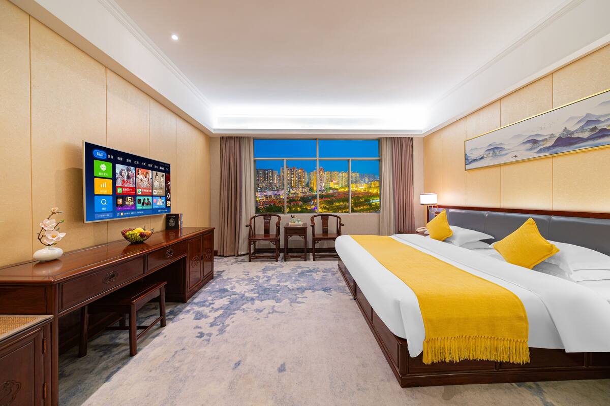 Una camera d'albergo con un letto grande, una scrivania con un computer, una TV a parete e una finestra con vista sulla città.