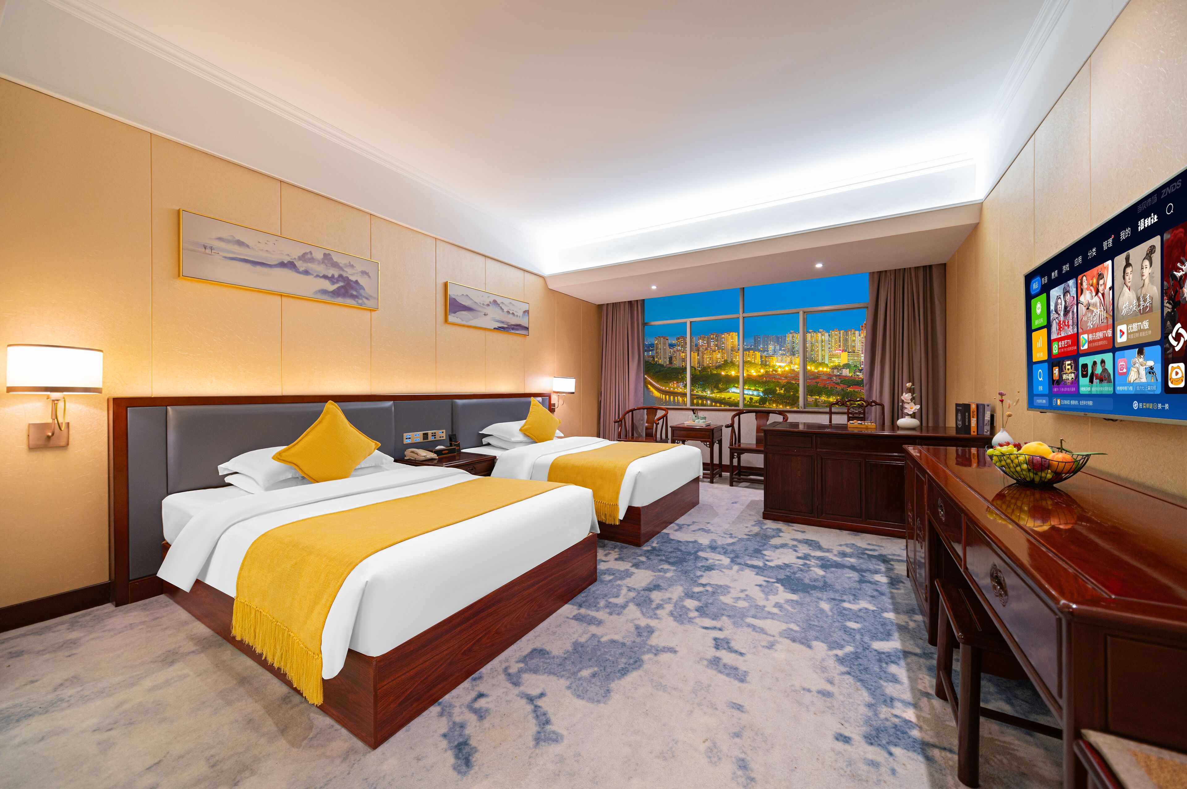 A hotel room with two beds, a desk, a chair, a TV, and a large window with a city view.