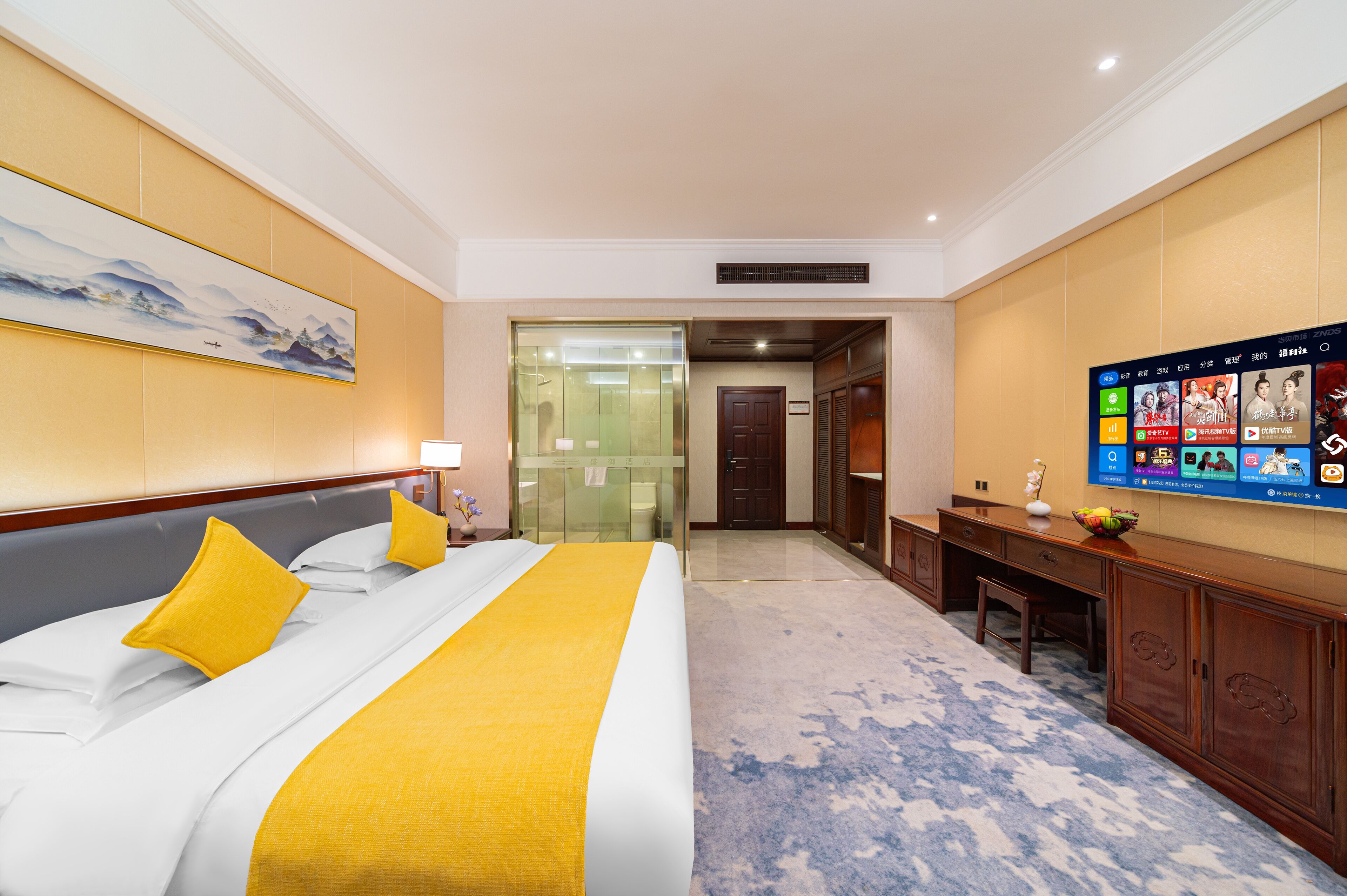 A hotel room with a large bed, a television mounted on the wall, a desk, and a door leading to another room.