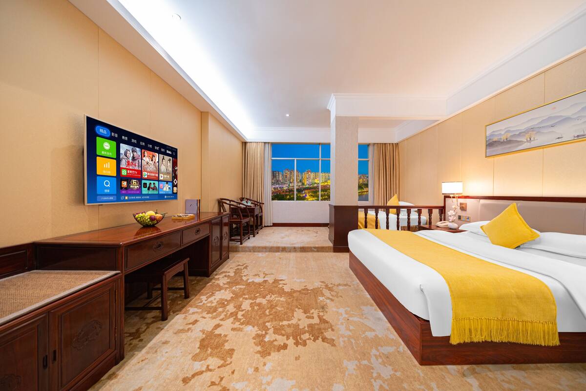 Una camera d'hotel con una grande TV a schermo piatto, un letto con una coperta gialla, una scrivania con una sedia e vista sulla città dalla finestra.