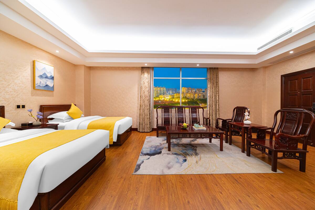Camera d'albergo con due letti, una zona salotto con tavolo e sedie, e un'ampia finestra che si affaccia sulla città.