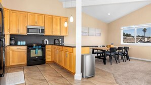 Une cuisine moderne avec des meubles en bois, un four à micro-ondes en acier inoxydable et un four noir. On y trouve un coin repas avec une table et des chaises, ainsi qu’une fenêtre donnant sur des maisons.