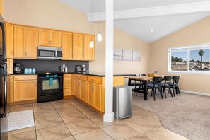 A modern kitchen with wooden cabinets, a stainless steel microwave, and a black oven. There is a dining area with a table and chairs, and a window with a view of houses.