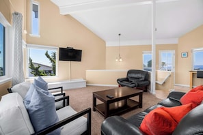 Una sala de estar con sofá, sillón y mesa de centro. Hay un televisor montado en la pared y una ventana con vistas al exterior.