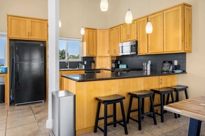 A modern kitchen with wooden cabinets, a black refrigerator, a stainless steel microwave, and a black countertop. There are three black stools and a wooden table.