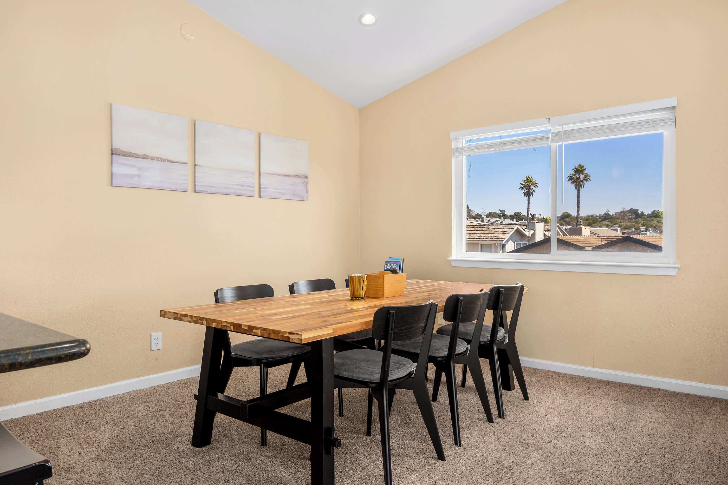 A room with a wooden table, six chairs, and a window with a view of palm trees and houses.