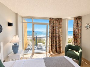 A hotel room with a bed, a chair, a table with a lamp, a view of the beach, and a balcony with curtains. - Ocean Breeze Hideaway (Myrtle Beach)