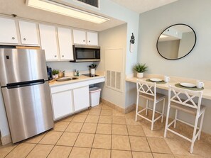 A modern kitchen with a stainless steel refrigerator, microwave, and white cabinets. There is a small bar area with two stools and a mirror on the wall. - Ocean Breeze Hideaway (Myrtle Beach)