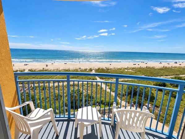 A balcony with white chairs and a view of a beach with people and the ocean.