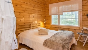 A cozy wooden cabin room with a bed, a window with blinds, a white robe, and a small table.