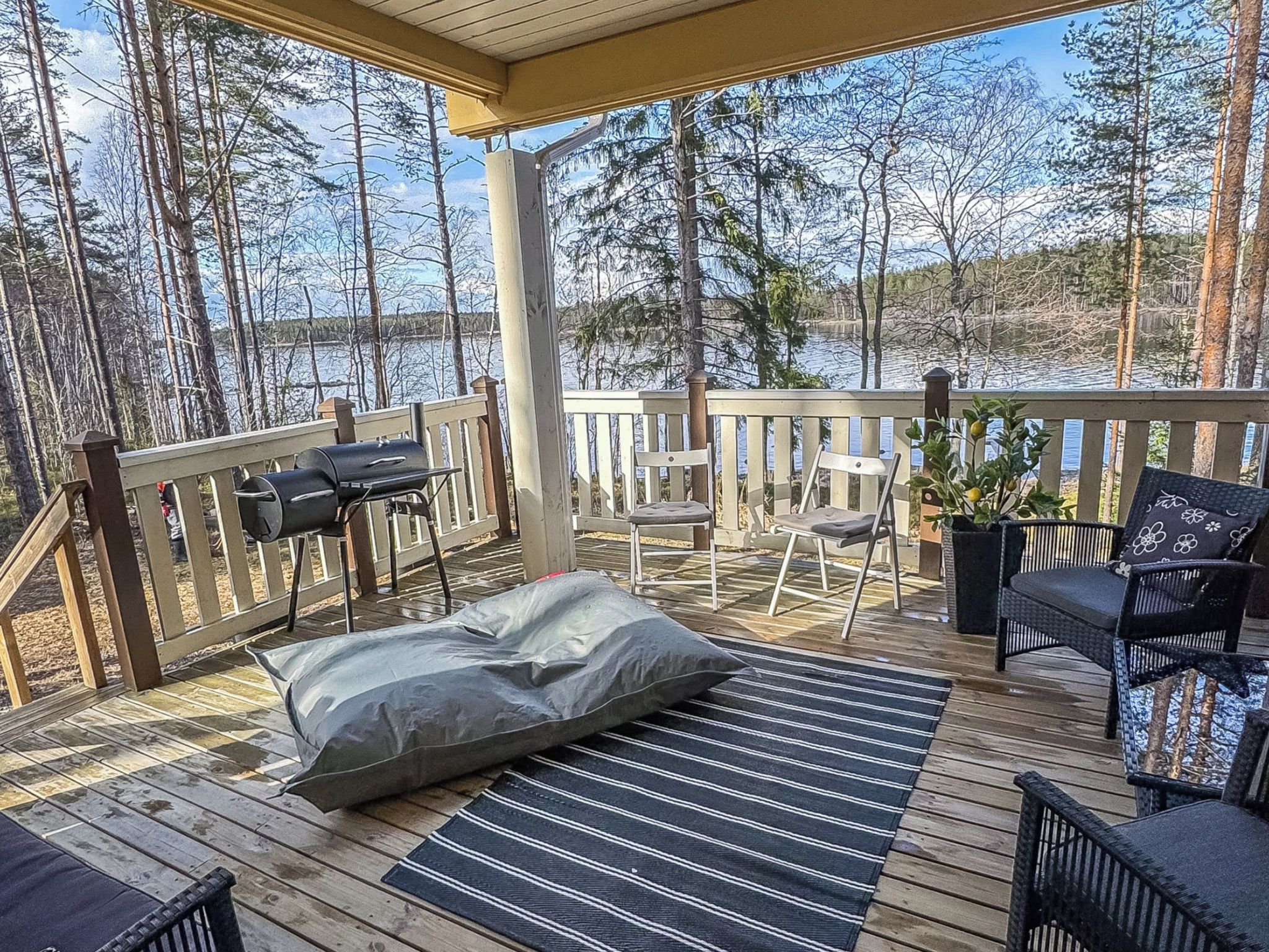 Une terrasse couverte, aménagée avec du mobilier d’extérieur, un barbecue et offrant une vue sur un lac entouré d’arbres.