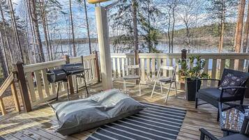 Une terrasse couverte, aménagée avec du mobilier d’extérieur, un barbecue et offrant une vue sur un lac entouré d’arbres.