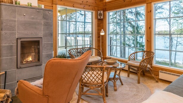 A cozy cabin interior with a fireplace, wicker furniture, and large windows offering a view of trees and a lake.