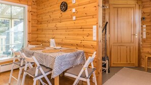 A wooden cabin interior with a dining table set for four, a clock on the wall, and a door leading outside.