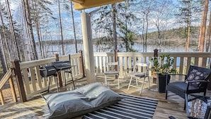 A wooden deck with a grill, chairs, and a view of a lake surrounded by trees.