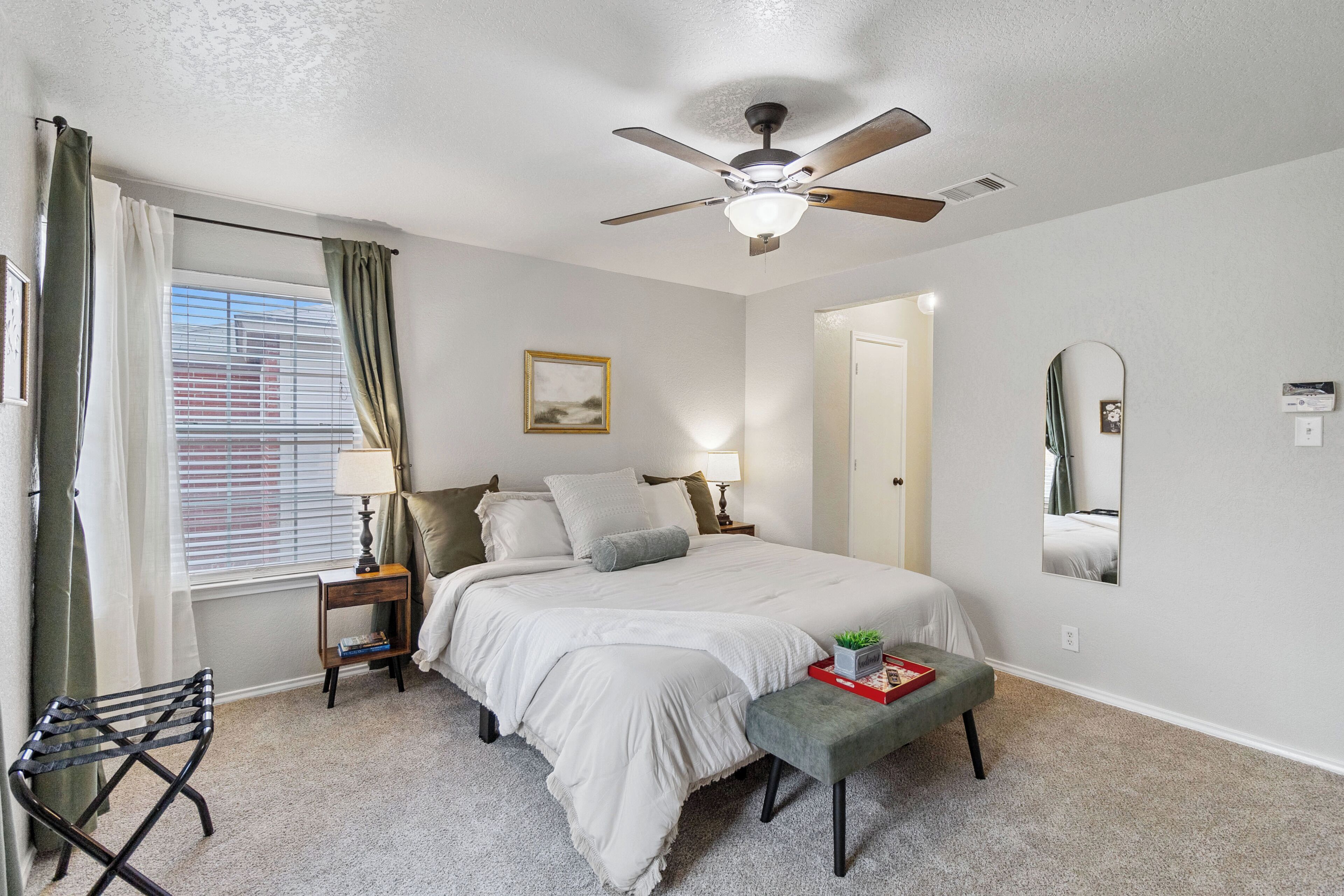 A bedroom with a bed, a ceiling fan, a nightstand, a lamp, a bench, a window with curtains, and a mirror on the wall.