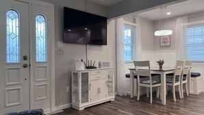 A dining area with a TV mounted above a white cabinet, a dining table with chairs, and a window with white curtains.