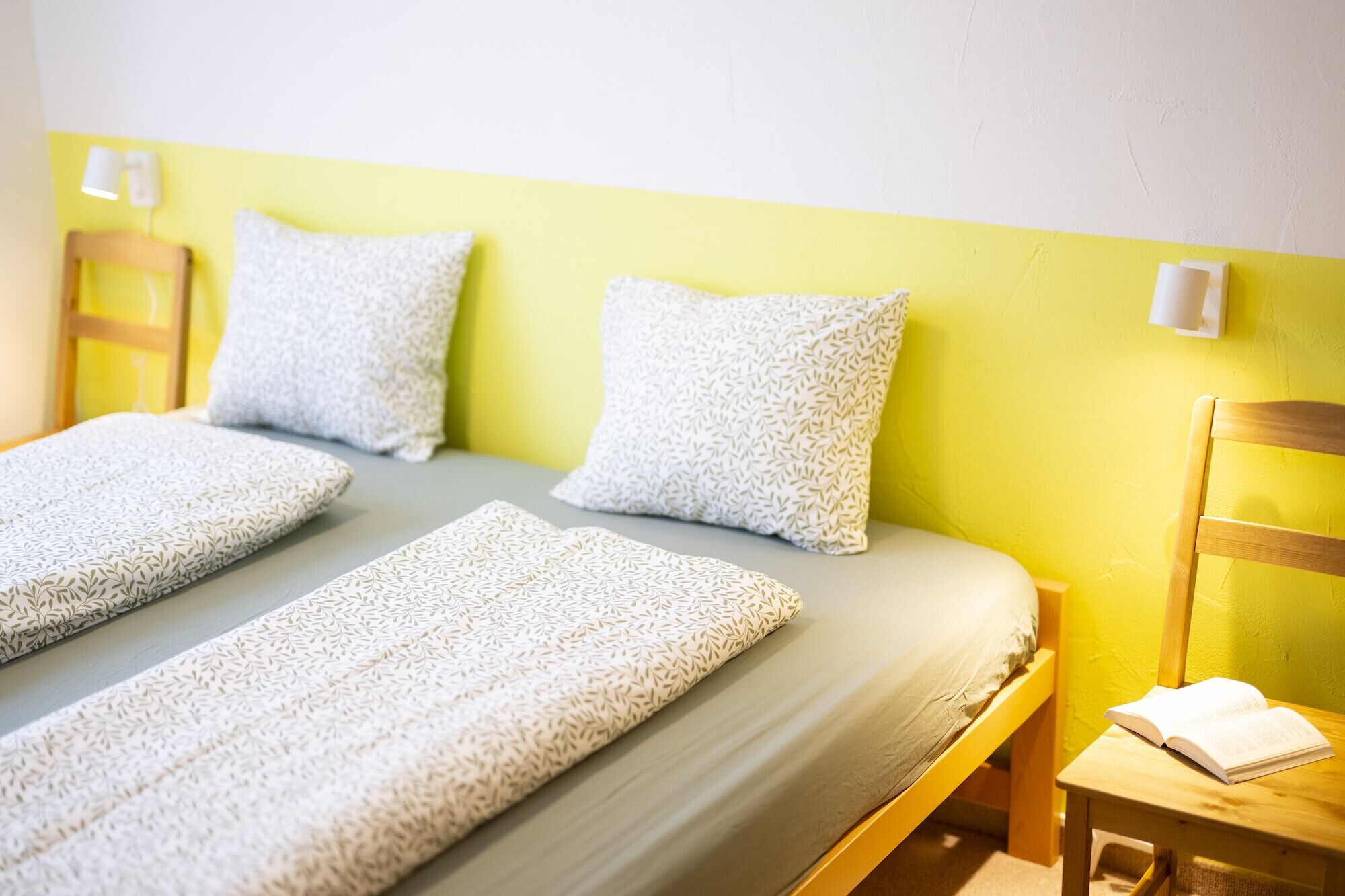 Una cama con dos almohadas y una colcha, una mesita de noche con un libro, y una silla.