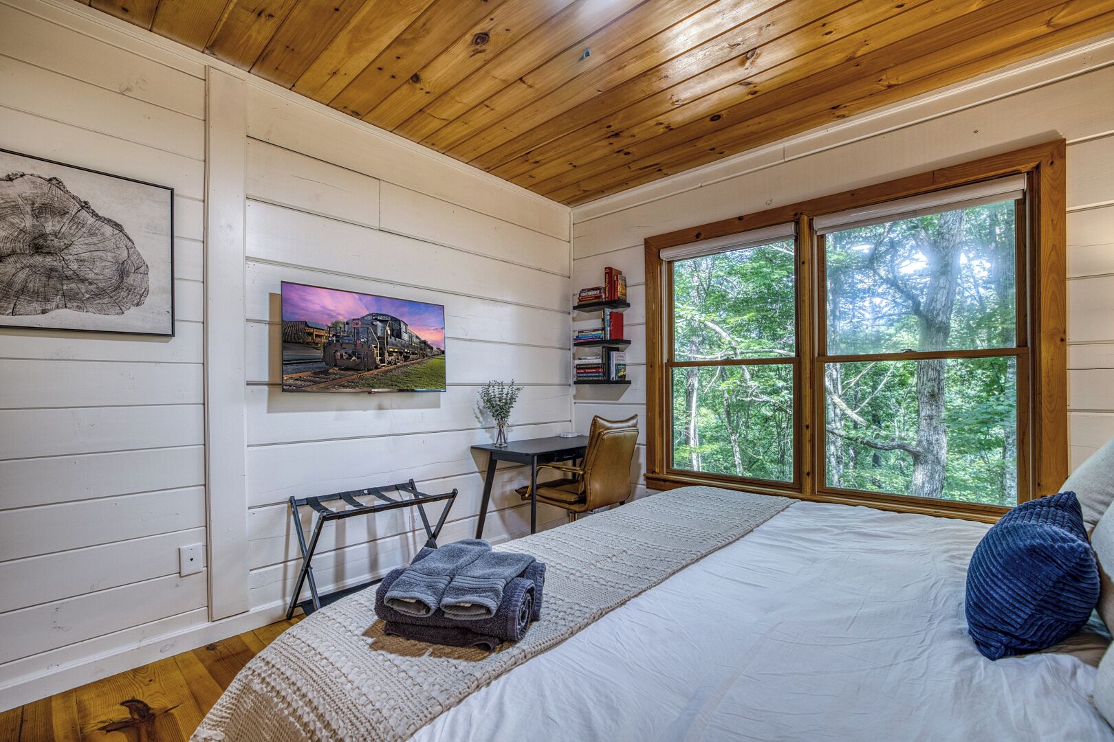 A bedroom with a large bed, a desk with a chair, a bookshelf, and a window with a forest view.