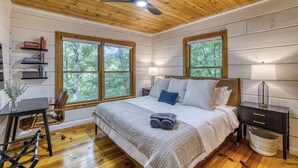 A bedroom with a wooden ceiling, a bed with pillows, a bedside table with a lamp, a nightstand, a bookshelf, and a window with a view of trees. - Dome, Hammock, Sauna, Golf, Best Location in BR (Blue Ridge)