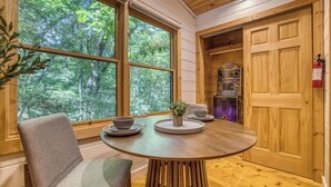 A cozy room with a wooden interior, a round table with a bowl and a plant, a chair, and a view of the forest through large windows. - Dome, Hammock, Sauna, Golf, Best Location in BR (Blue Ridge)